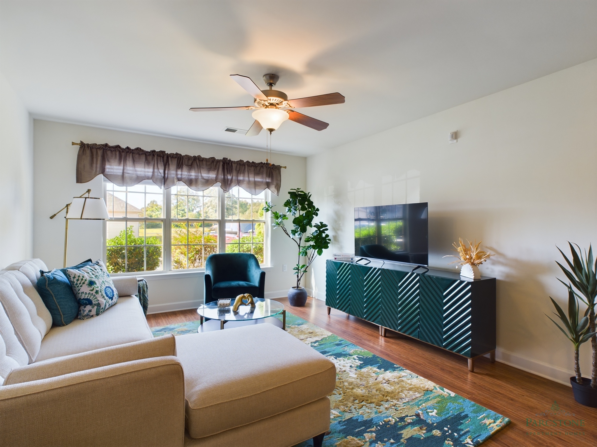 Apartments in Fayetteville, North Carolina A cozy living room at Parcstone Apartments features a ceiling fan, large window, couch, armchair, TV on a console, coffee table, potted plants, and a rug on wooden flooring.